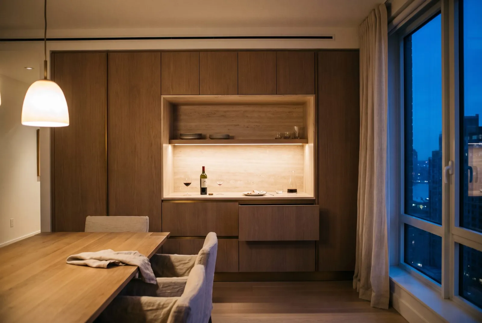 A brown oak storage wall with a lit travertine service niche, dining table in front